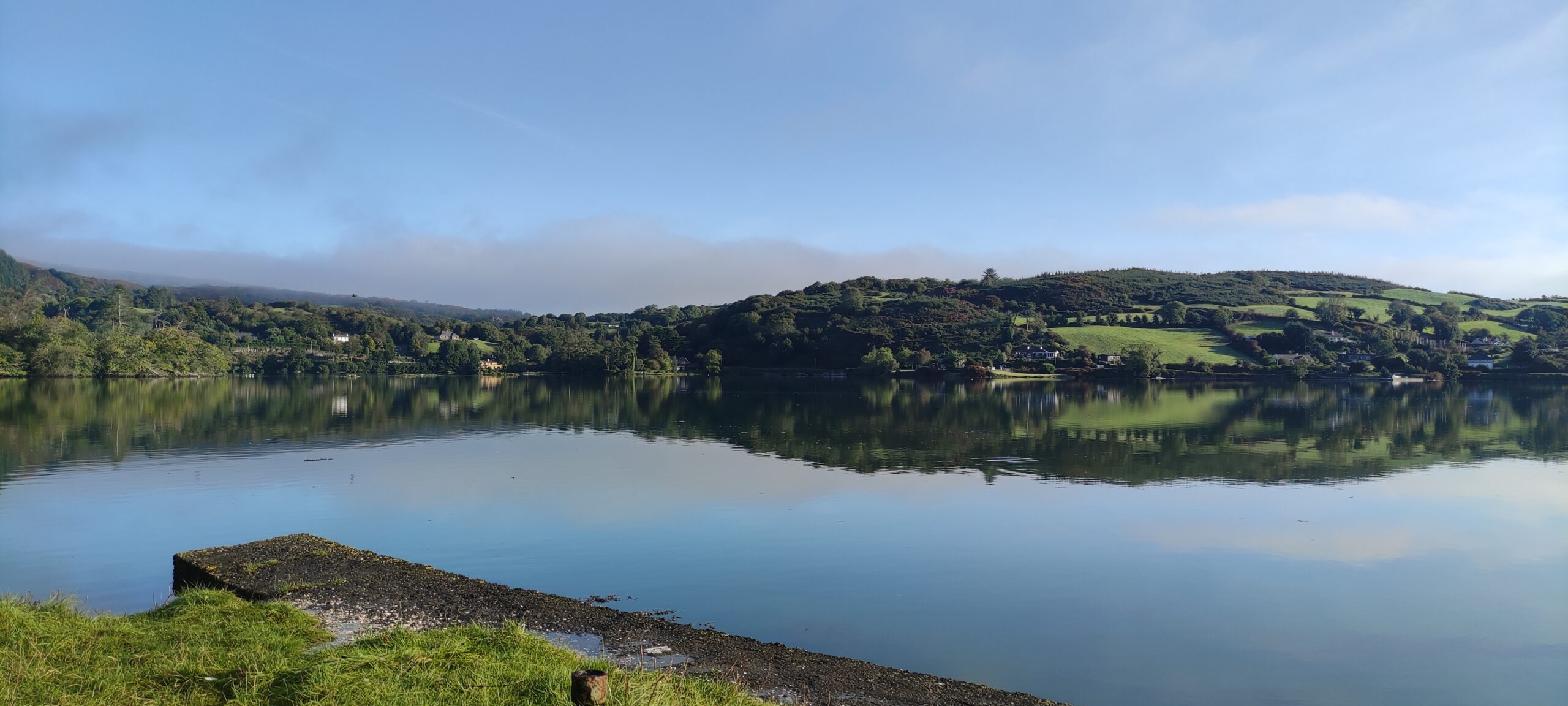 A photograph of a bright grey-blue lake with gentle deep green hills rolling in the background. The lake is reflecting the bright light blue sky which can be seen in the background. There is a small green verge in the very foreground of the photograph.